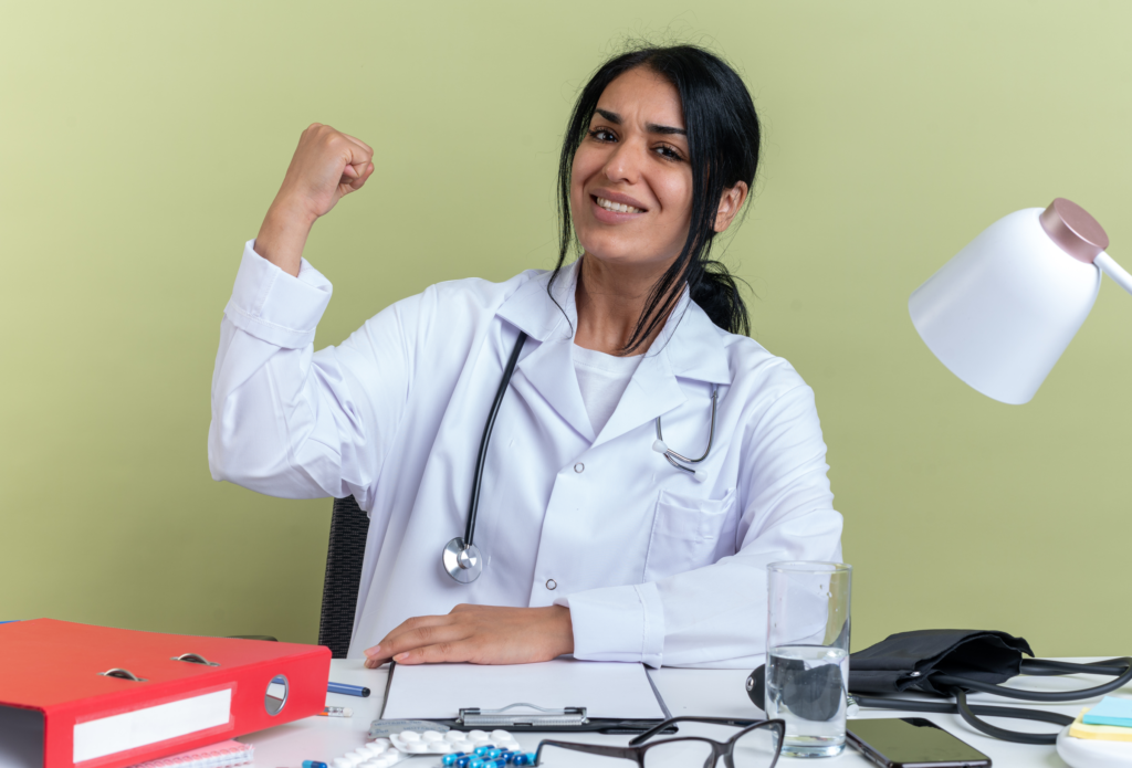smiling-young-female-doctor-wearing-medical-robe-with-stethoscope-sits-desk-with-medical-tools-showing-yes-gesture-isolated-olive-green-background